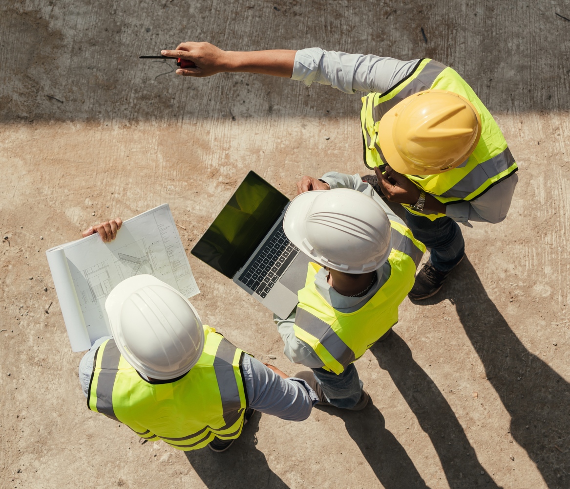 Three men, at a construction site, pointing, holding laptops and plans.