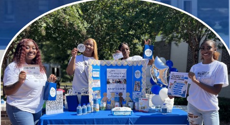 Four Fayetteville State University students standing behind a blue-draped table at an outdoor FSU Food Resource Pantry giveaway event, holding informational cards and tote bags.