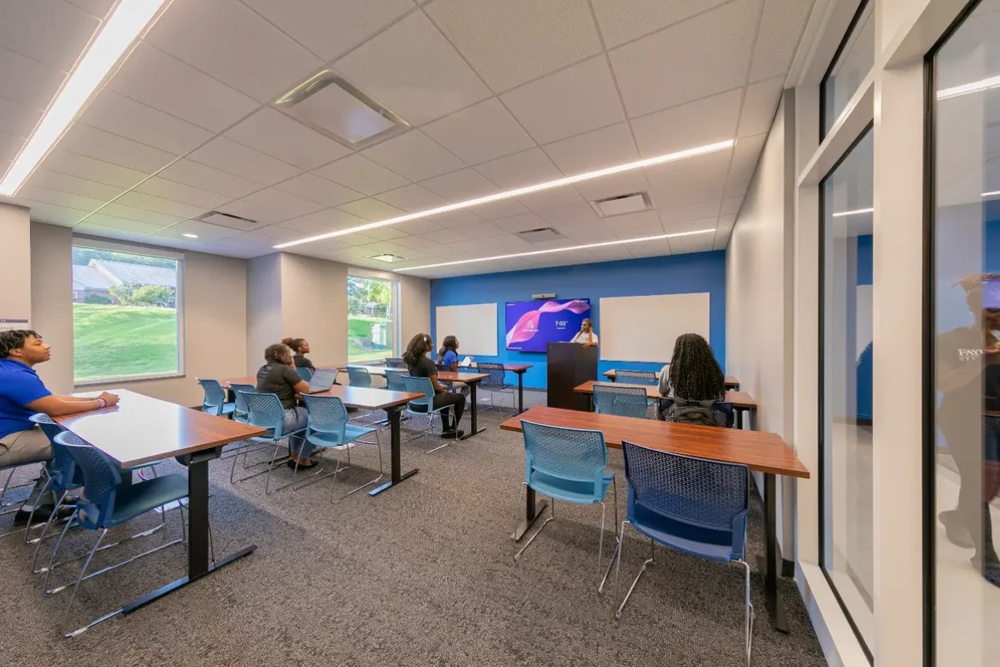 The first floor conference room in Bronco Pride Hall with serveral students seated at tables facing a presenter, whiteboards, and a presentation screen a the front of the room.