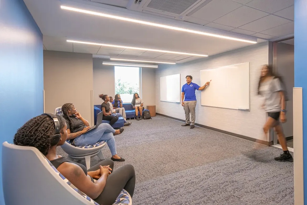 A hallway nook in Bronco Pride Hall serves as an informal gathering and learning space, with students seated in lounge chairs while a staff member presents at a whiteboard.