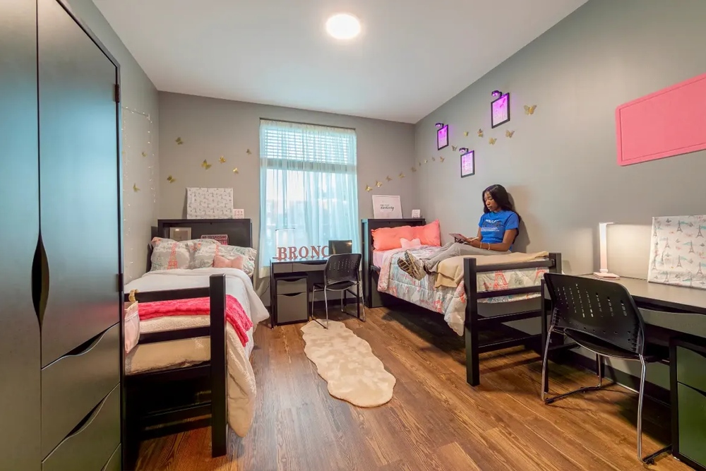 A furnished Bronco Pride Hall demo room with two beds, desks, chairs, and wardrobes. The room featurs wood-style flooring, blue and pink accents, decorative wall art, and a student seated on one of the beds.