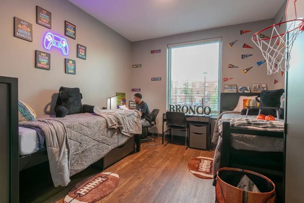A furnished Bronco Pride Hall demo room with two beds, desks, chairs, and storage furniture. The room features wood-style flooring with sports and gaming themed decor, including football rugs, pennants, and a wall-mounted basketball hoop. A student is stead at on of the desk.