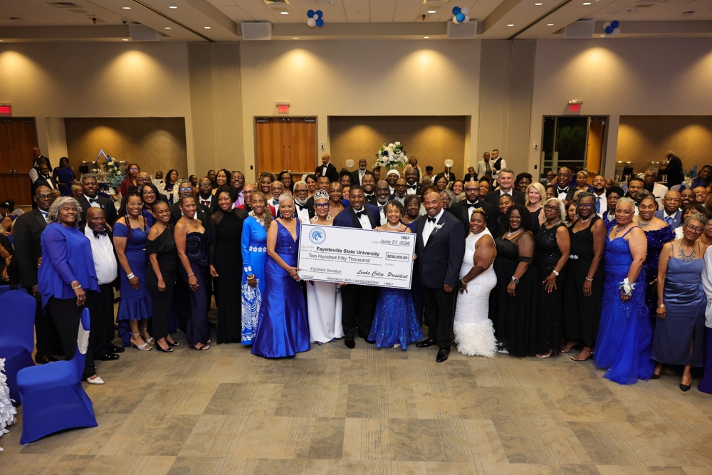 A large crowd gathered around Chancelor Adilson, all smiling as he and a few others hold a large white and blue novelty check.