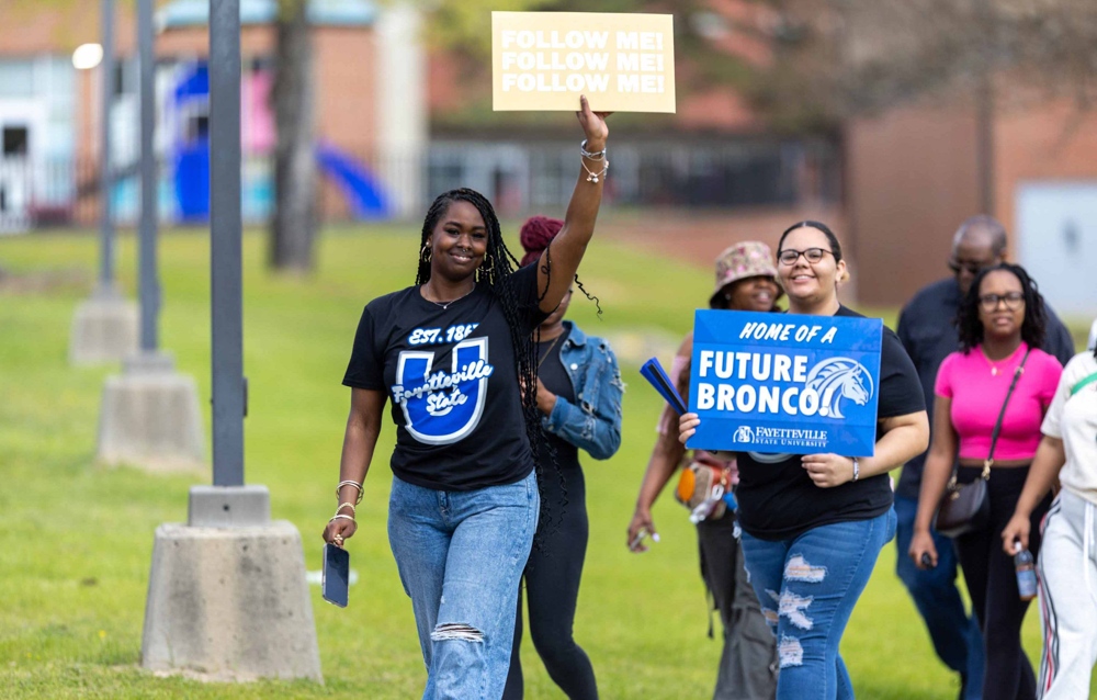 Two older students holding signs, one reads: Follow Me!; The other reads: Home of a Future Bronco