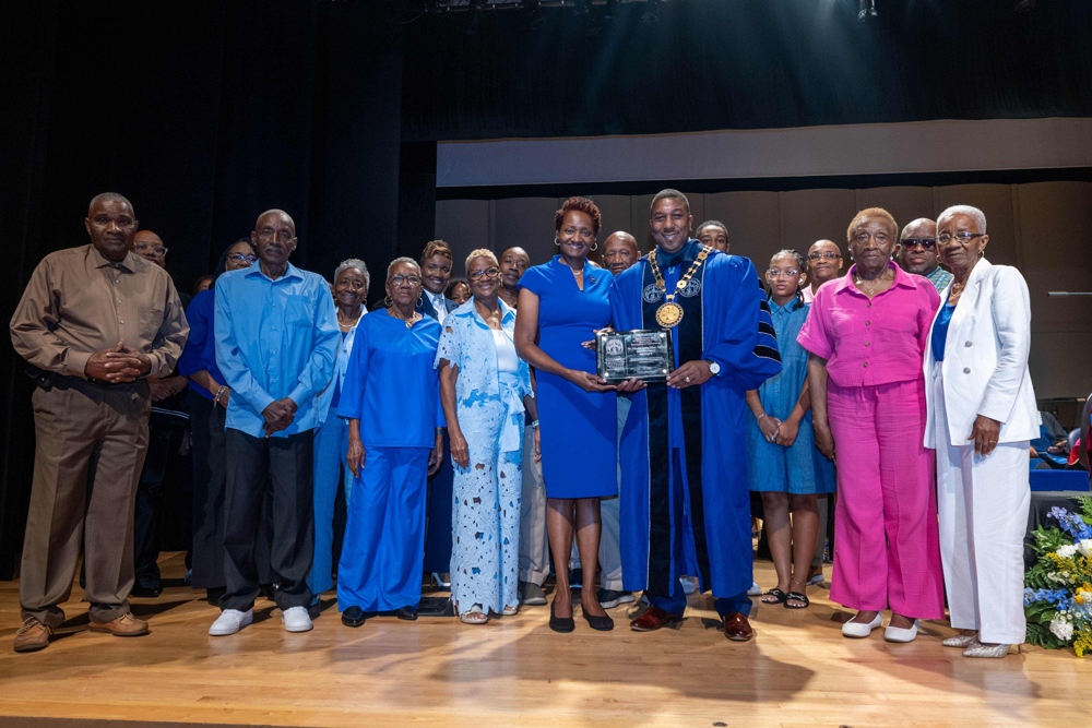 A number of smiling people fill the stage; up front the Chancellor is handing an award to a proud woman in a blue dress.