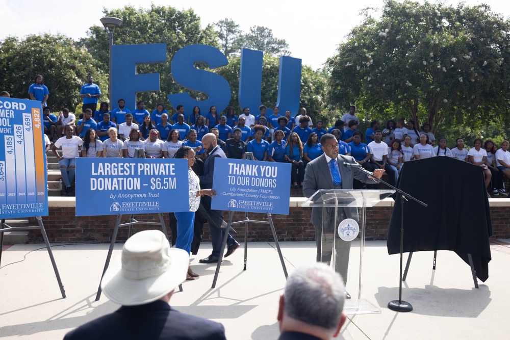 The Chancellor speaking at a podium, blue signs proclaiming donations can be seen.