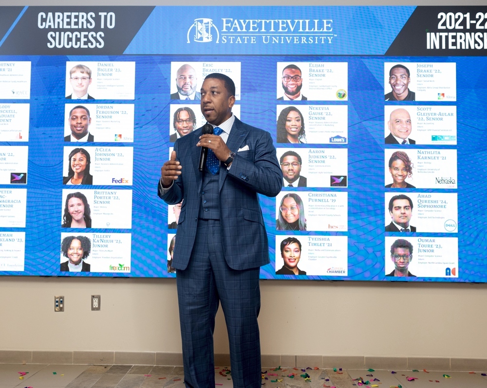 The chancellor standing with a mic, behind him is a presentation featuring the portraits of many students.